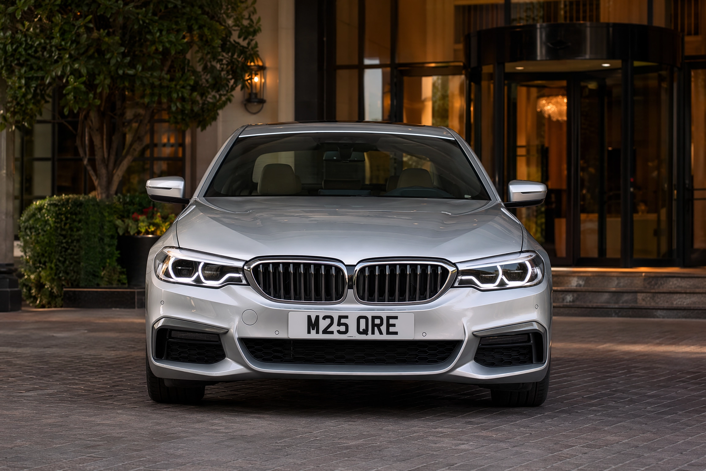 1125 M25 Chauffeurs Ltd Professional Chauffeur Service United Kingdom Front view of a silver BMW SUV parked in front of a glass building entrance, headlights on, license plate M25 QRE.