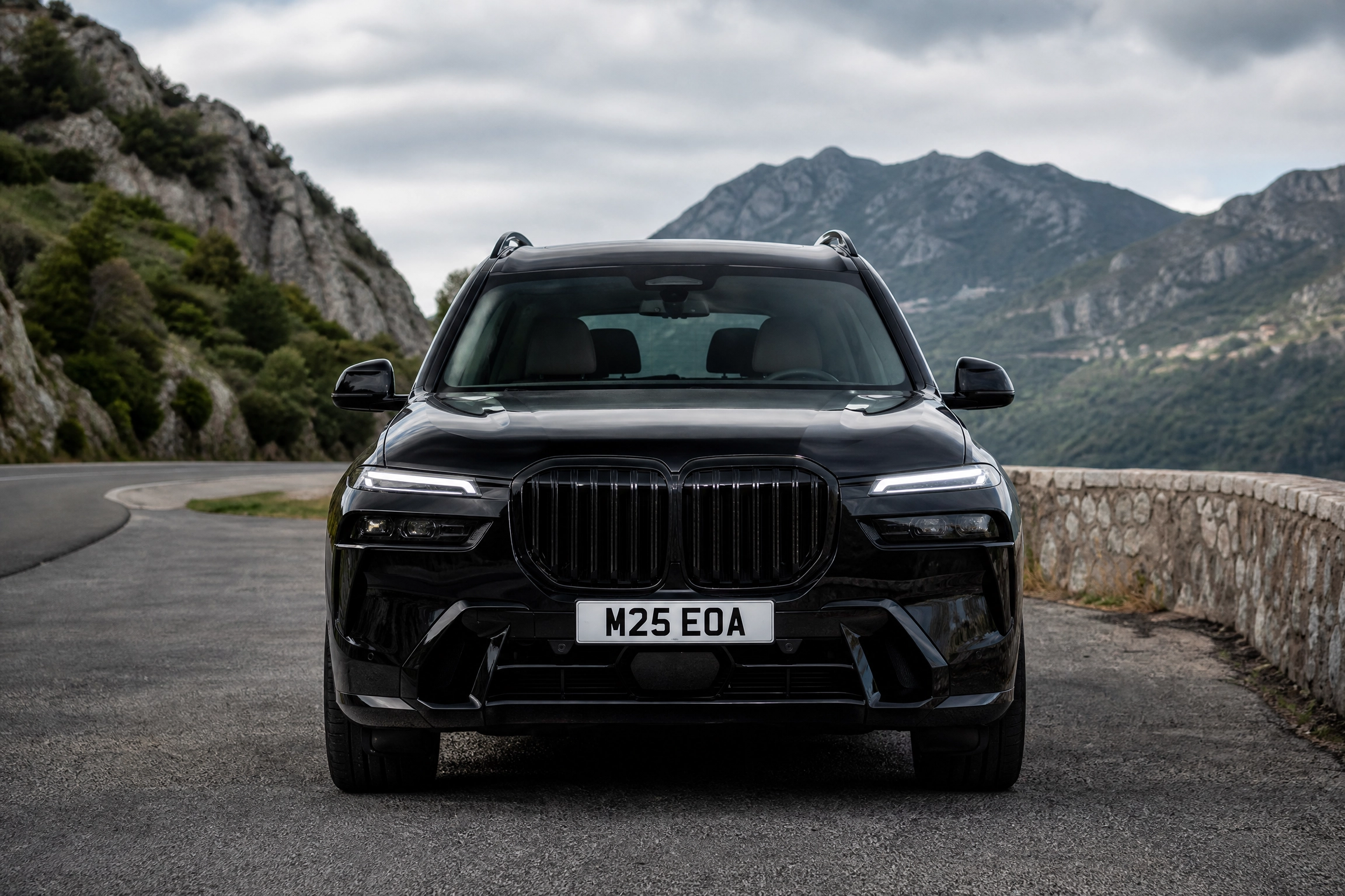 Front view of a black SUV on a winding mountain road, rocky hillside to the left and distant mountains in the background.