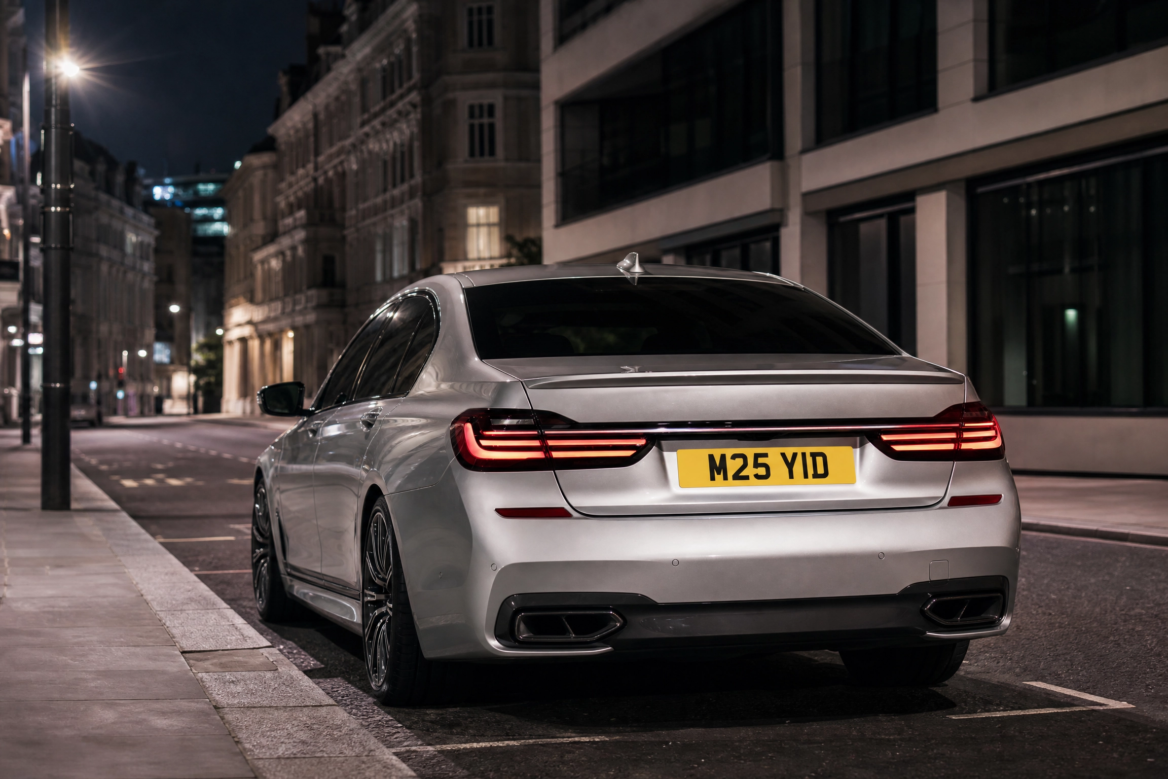 Rear view of a silver luxury sedan parked on a city street at night, with illuminated taillights and the license plate M25 YID.