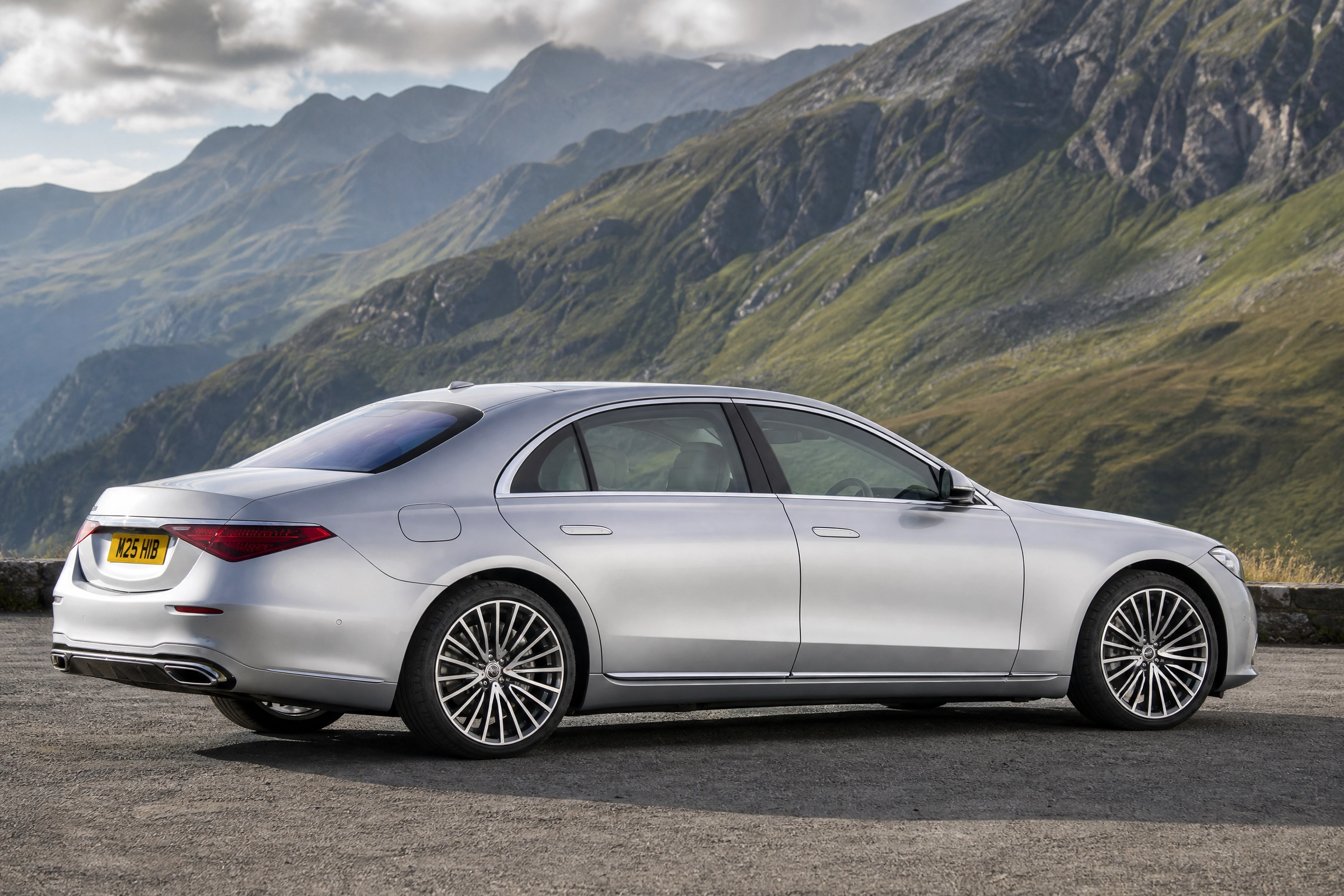 Chauffeur Hire in Yate by M25 Chauffeurs Ltd 1 M25 Chauffeurs Ltd Professional Chauffeur Service United Kingdom Silver luxury sedan parked on a mountain road with rugged green slopes and cloudy sky in the background; visible UK-style yellow license plate M25 HIB on rear.