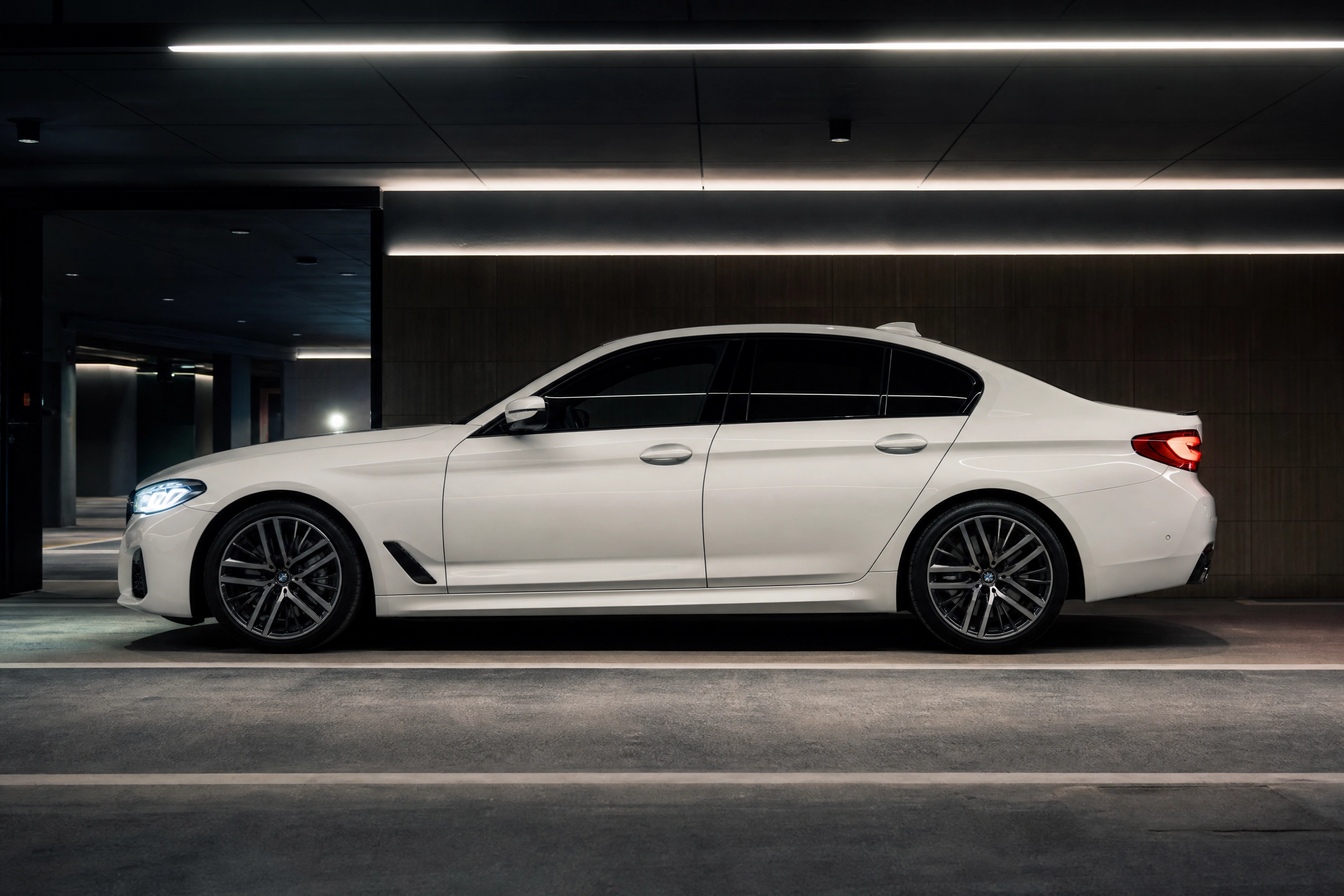 Chauffeur Hire in Yate by M25 Chauffeurs Ltd M25 Chauffeurs Ltd Professional Chauffeur Service United Kingdom White luxury sedan parked in a dim, modern underground garage with linear ceiling lights overhead.