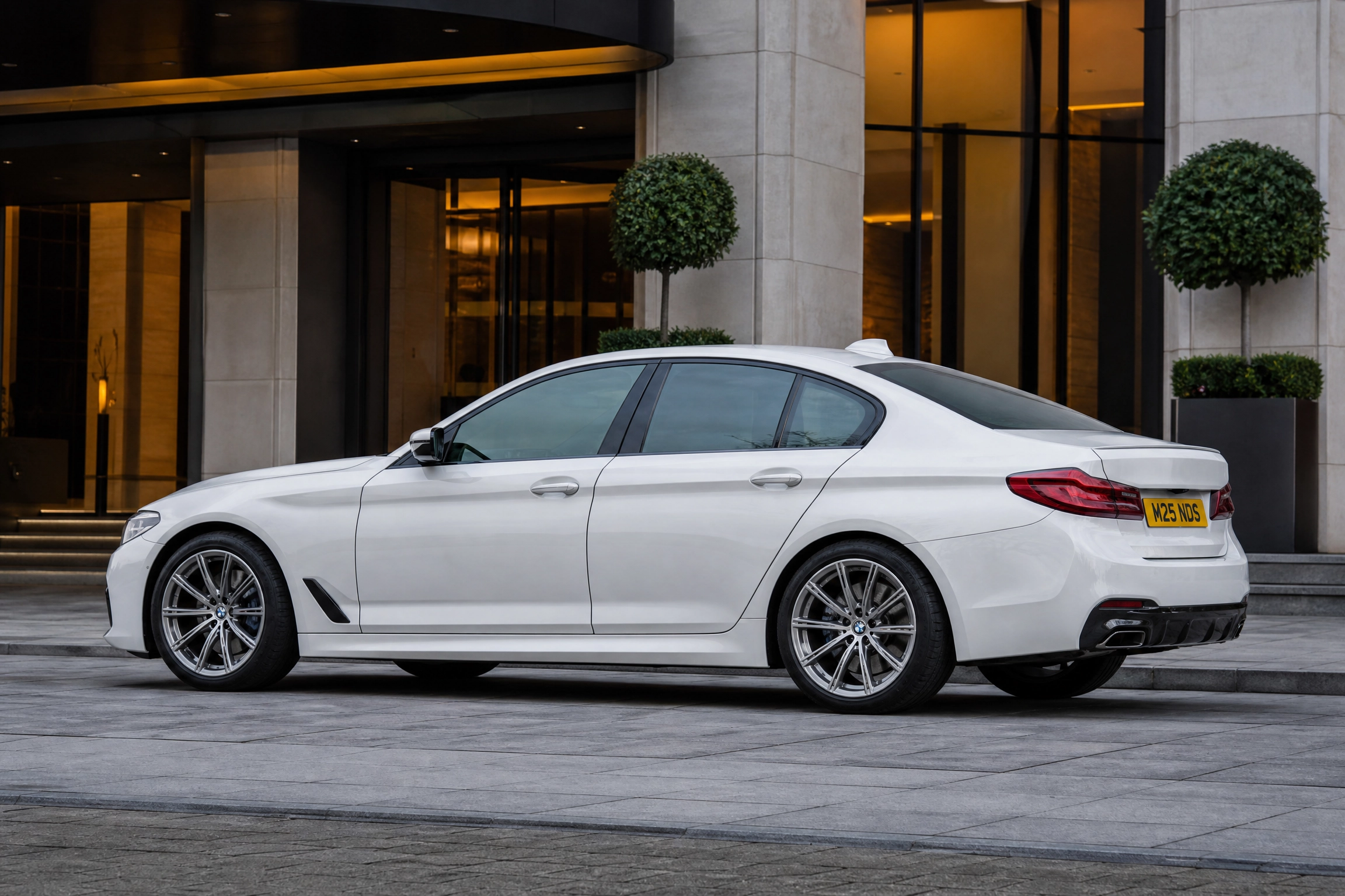Chauffeur Service in Thorne by M25 Chauffeurs Ltd 1 M25 Chauffeurs Ltd Professional Chauffeur Service United Kingdom White BMW sedan parked in front of a modern building with gold-lit windows and trimmed trees in planters; tail light visible on right.